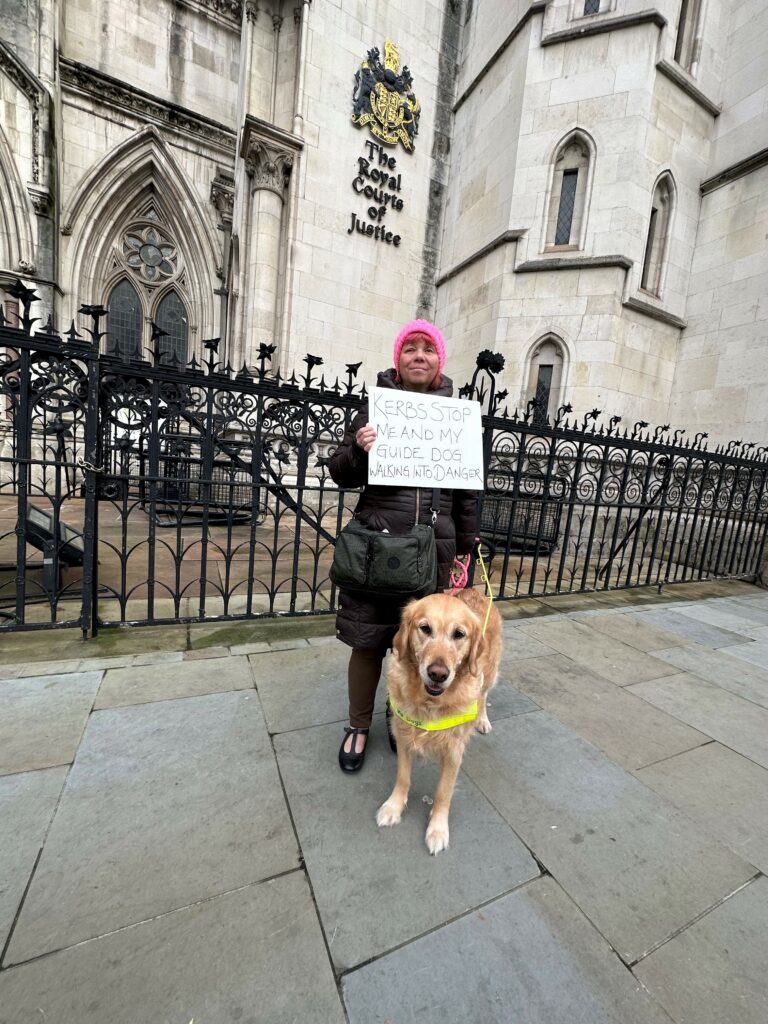 Sarah Leadbetter outside the Royal Courts of Justice in London, with her Guide Dog Nellie, holding a sign that states ‘Kerbs Stop Me and My Guide Dog Walking Into Danger’