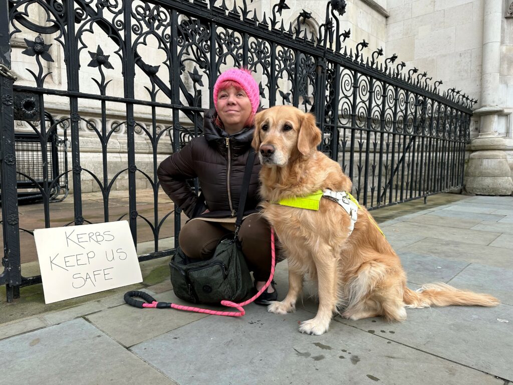 Sarah Leadbetter outside the Royal Courts of Justice in London, with her Guide Dog Nellie, holding a sign that states ‘Kerbs Kept Us Safe'