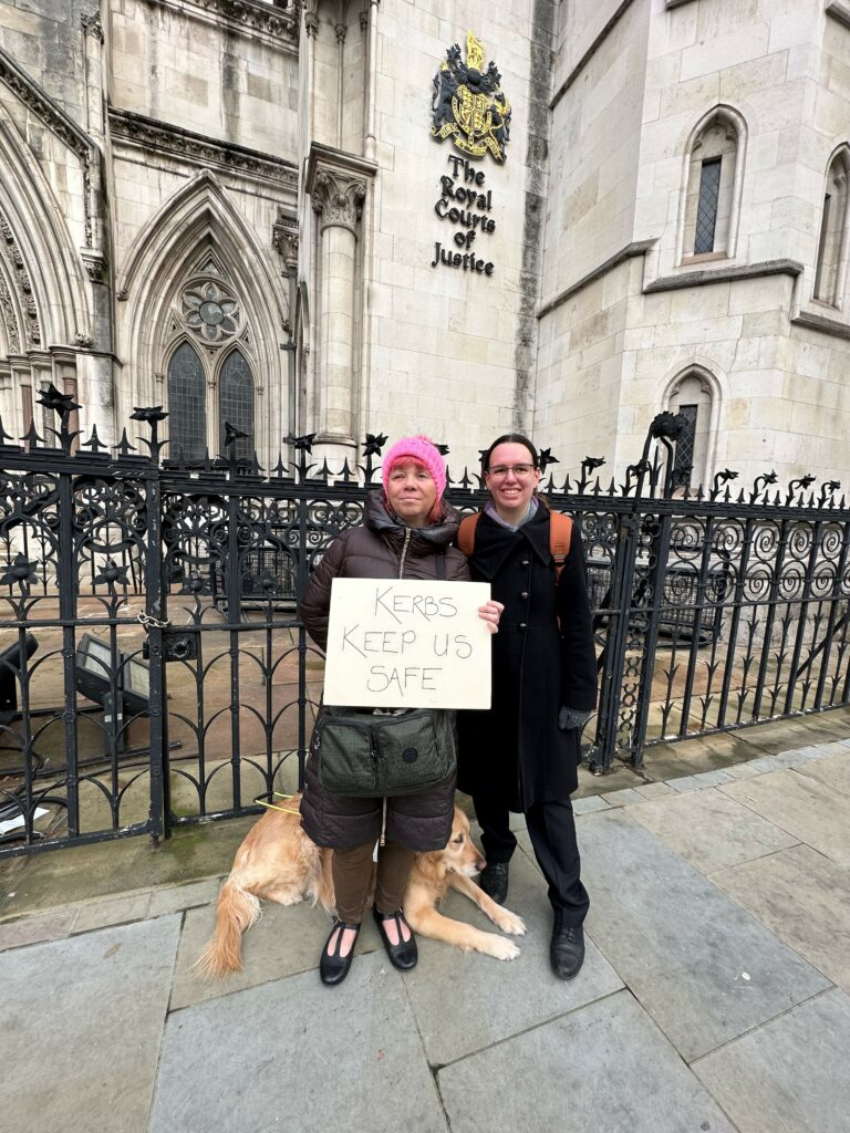 Sarah Leadbetter outside the Royal Courts of Justice in London, with her Guide Dog Nellie and Elizabeth Cleaver from Bindmans LLP holding a sign that states ‘Kerbs Kept Us Safe'