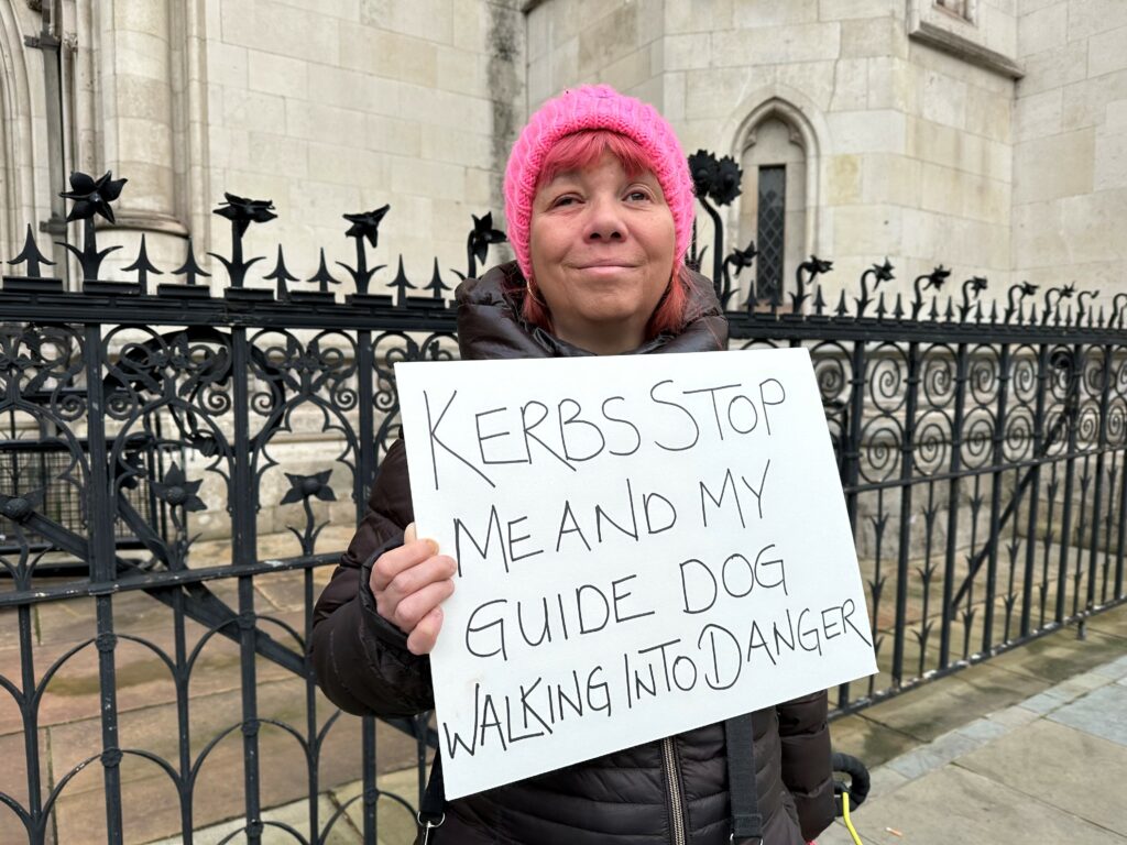 Sarah Leadbetter outside the Royal Courts of Justice in London, holding a sign which states ‘Kerbs Stop Me and My Guide Dog Walking Into Danger’